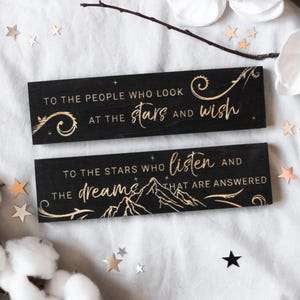 Op de afbeelding: Twee zwarte houten platen met gouden letters. De bovenste plaat leest "TO THE PEOPLE WHO LOOK AT THE stars AND wish." De onderste plaat leest "TO THE STARS WHO listen AND THE dreams THAT ARE ANSWERED."