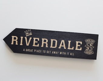 May include: Black wooden sign with a white arrow pointing to the right. The sign says "Riverdale" in white letters with a crown above the word and "A great place to get away with it all" below. On the right side of the sign is a black and white logo with a snake and the words "South Side Serpents".