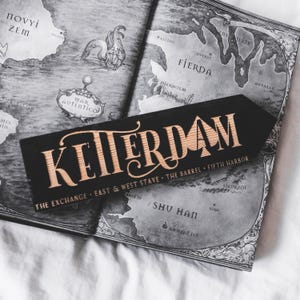 Peut inclure: Un panneau en bois noir avec le mot "KETTERDAM" en lettres dorées stylisées, posé sur une carte ouverte. Le panneau comprend également le texte "THE EXCHANGE • EAST & WEST STAVE • THE BARREL • FIFTH HARBOR". La carte est en niveaux de gris.