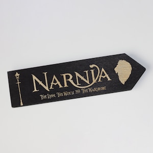 Pode incluir: Marca-páginas de madeira preta com letras douradas que dizem "Narnia O Leão, a Feiticeira e o Guarda-Roupa". Uma cabeça de leão dourada está no lado direito do marca-páginas.