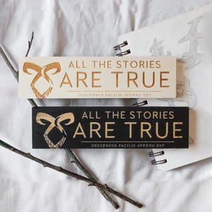 Könnte beinhalten: Zwei rechteckige Holzschilder mit dem Text "ALL THE STORIES ARE TRUE" und "DESCENSUS FACILIS AVERNO EST". Eines ist hell mit braunem Text, das andere ist schwarz mit goldenem Text. Auf der linken Seite befindet sich ein dekoratives Bogendesign.