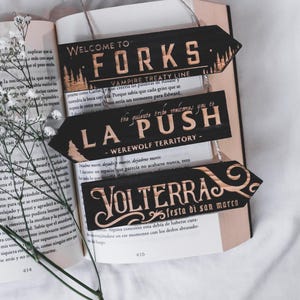 May include: Three black wooden arrow signs with gold lettering and designs. The signs read "Welcome to Forks - Vampire Treaty Line", "La Push - Werewolf Territory", and "Volterra - Festa di San Marco". The signs are hanging over an open book.