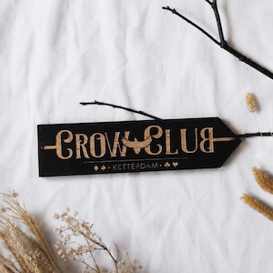 Pode incluir: Uma placa de madeira preta com as palavras "CROW CLUB" em uma fonte estilizada, com um gráfico de pássaro. A placa também inclui o texto "KETTERDAM" e elementos decorativos. A placa está em um fundo branco com plantas e galhos secos.