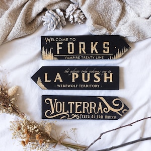 Puede incluir: Tres letreros de flecha de madera negra con letras doradas. Los letreros dicen "Welcome to Forks, Vampire Treaty Line", "La Push, Werewolf Territory" y "Volterra, festa di san marco". Los letreros están dispuestos sobre una superficie blanca con flores secas y una tela beige.