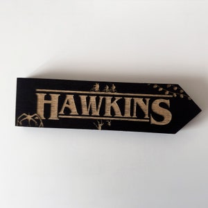 May include: Black wooden sign with the word "HAWKINS" in brown lettering. The sign is shaped like an arrow pointing to the right. There are small images of figures on bicycles and a monster on the sign.