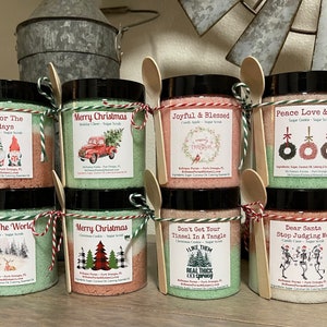 May include: A collection of Christmas-themed sugar scrubs in clear jars, each with a wooden spoon and tied with twine. Labels feature festive designs like gnomes, wreaths, and a red truck. The jars are filled with colorful, layered scrubs.