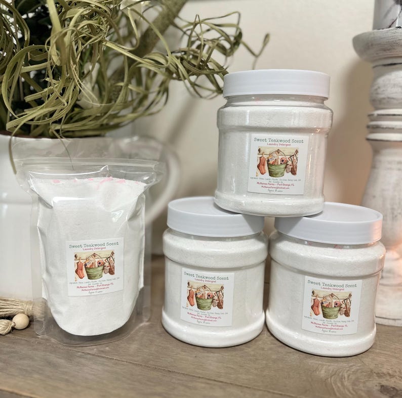 Op de afbeelding: Vier witte plastic containers met wasmiddel met een Sweet Teawood geurlabel. Het label heeft een vintage illustratie van een waslijn met hangende was.