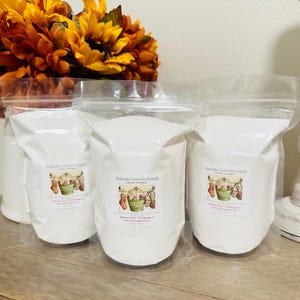 Op de afbeelding: Drie witte plastic zakken gevuld met wasmiddel. De zakken zijn gelabeld met "Autumn Leaves Scent Laundry Detergent". De zakken staan op een houten oppervlak.