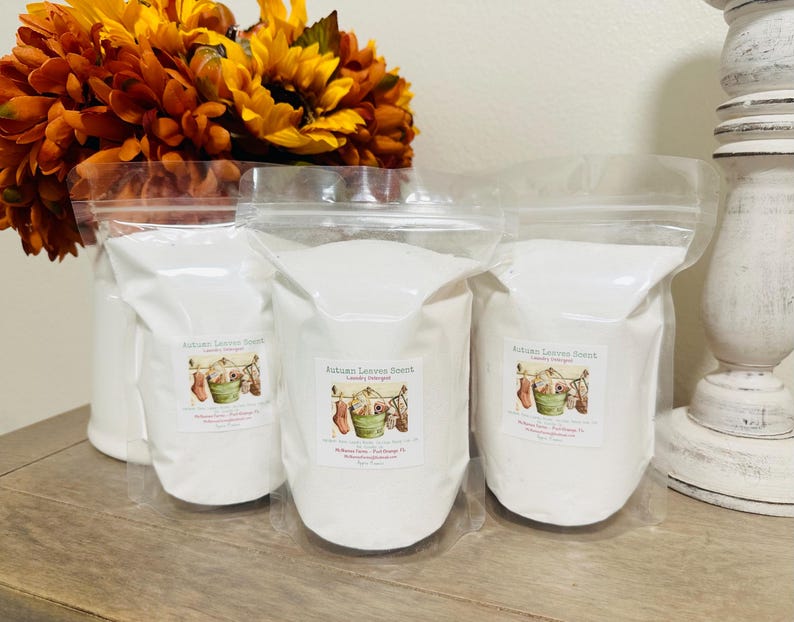 Op de afbeelding: Drie witte plastic zakken gevuld met wasmiddel. De zakken zijn gelabeld met "Autumn Leaves Scent Laundry Detergent". De zakken staan op een houten oppervlak.