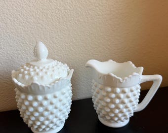 May include: A white milk glass sugar bowl with a matching lid and a creamer pitcher. Both pieces feature a raised hobnail texture, a scalloped rim, and a pedestal base. The sugar bowl lid is topped with a pointed, textured finial.