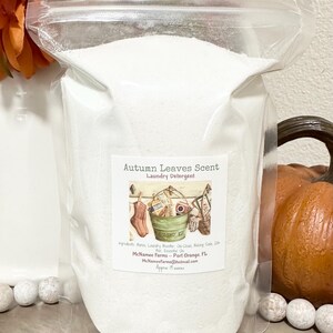 Op de afbeelding: Een witte hersluitbare zak gevuld met wasmiddelpoeder. Het etiket luidt "Autumn Leaves Scent Laundry Detergent" met een tekening van een wasmand en een paar sokken. Het etiket bevat ook de ingredi&euml;nten, de bedrijfsnaam "McNamee Farms", de locatie "Port Orange, FL", het e-mailadres "McNameeFarms@hotmail.com" en de grootte "Approx 19 ounces".