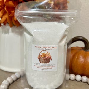 Op de afbeelding: Een witte hersluitbare plastic zak gevuld met wit wasmiddelpoeder. De zak heeft een label met de tekst "Sweet Pumpkin Scent Laundry Detergent" met een mand vol herfstbladeren en de tekst "Melance Farms - Port Orange, FL MelanceFaras@hotmail.com".