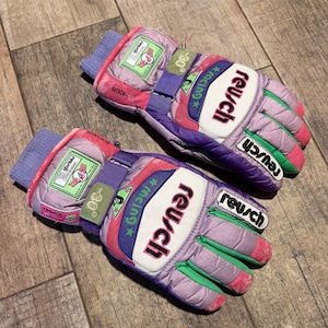 Op de afbeelding: Een paar Reusch racehandschoenen in lavendel, roze, groen en wit. De handschoenen zijn voorzien van paarse verstelbare polsbanden, groene patches met de tekst -30 graden en witte knokkelbeschermers met de zwarte tekst Reusch racing.