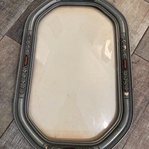 May include: An oval-shaped picture frame with a metallic silver finish and a smooth, rounded border. The frame features four decorative rectangular insets, each containing a polished amber-colored stone accent set against a textured silver base.