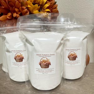 Op de afbeelding: Drie witte plastic zakken gevuld met wasmiddel. Elke zak heeft een label met de tekst "Sweet Pumpkin Scent Laundry Detergent". De zakken liggen op een houten oppervlak.
