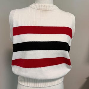 Op de afbeelding: Een mouwloos gebreid vest met een geribbelde ronde hals en zoom. Het kledingstuk is voornamelijk wit met horizontale strepen in rood en zwart over de borst en het midden, gemaakt van een zachte, getextureerde gebreide stof.