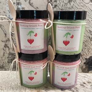Op de afbeelding: Vier glazen potten met suikerscrub met feestelijke Grinch-thema labels. De potten zijn vastgebonden met touw en hebben houten lepels bevestigd. De labels hebben een rood hart, een groene Grinch-hand en tekst die "Resting Grinch Face" en "I Am 100% That Grinch" zegt.