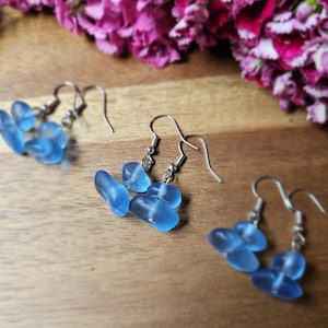 May include: Three pairs of drop earrings featuring smooth, translucent light blue sea glass pieces suspended from silver-toned metal hooks. Each earring consists of two rounded, pebble-like glass shapes linked together in a vertical arrangement.