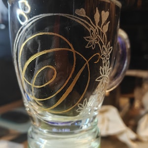 May include: A clear glass mug featuring a hand-painted gold spiral design on the front, complemented by a delicate white etched floral pattern along the side. The mug has a sturdy handle and a smooth, transparent finish, showcasing the contrast between the metallic gold paint and the fine white line work.