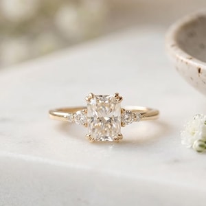 Könnte beinhalten: Ein goldener Ring mit einem großen, rechteckigen, klaren Edelstein als Mittelstück. Kleinere, klare Edelsteine flankieren den Hauptstein. Der Ring ist vor einem weißen Hintergrund mit unscharfen Blumen und einer dekorativen Schale platziert.