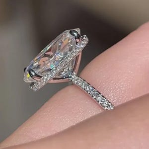 Op de afbeelding: Een close-up van een diamanten ring met een grote, heldere, peervormige steen. De ring heeft een halo van kleinere diamanten rond de hoofdsteen en langs de band. De ring is gezet in een zilverkleurig metaal.