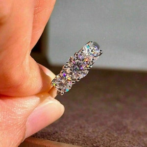 May include: A silver ring with three large, round, clear gemstones. The gemstones are set close together, and the ring is held between fingers. The gemstones sparkle and reflect light, creating a brilliant display.