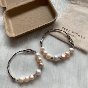 May include: Two silver bracelets with a delicate, irregular design. Each bracelet is adorned with a row of white and pink pearls.