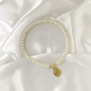 May include: A white pearl necklace with a gold coin pendant. The necklace is on a white satin background.