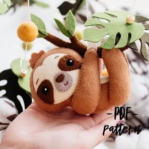 May include: A brown felt sloth hanging from a green felt branch with a green felt monstera leaf. The sloth has a white face with black eyes and a brown nose. The text "PDF Pattern" is written in black on a white background.