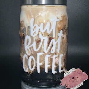 May include: A clear glass tumbler with a silver lid and straw, filled with a coffee-like beverage. The words "but first COFFEE" are in white script on the front. The drink has a marbled appearance with shades of brown and cream.