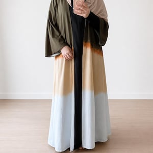 May include: A long, open-front abaya featuring a colour-blocked design with horizontal bands of olive green, tan, and light blue. The garment has wide, loose sleeves and is worn over a solid black floor-length dress, paired with a beige headscarf.