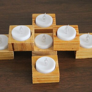 May include: A decorative arrangement of seven battery-operated LED tea lights. Each white candle is set in a small, square, natural wood holder. The holders are arranged in a cross shape on a dark brown surface.