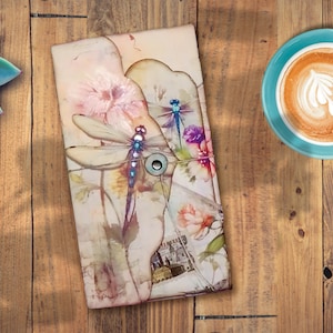 May include: A watercolor-style journal cover with a dragonfly design. The cover features a light brown background with pink, purple, and blue flowers and dragonflies. The journal is closed and has a metal button closure.