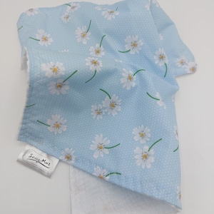 May include: A light blue fabric featuring a white daisy print and white polka dots. The fabric is folded over a white, textured material. A tag with the text "Snug Muz" is attached to the fabric.