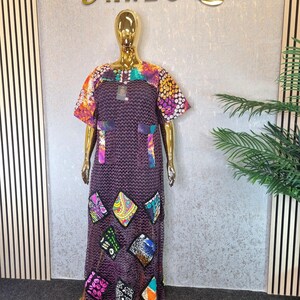 May include: A long, dark purple dress with short sleeves and colourful patterned accents. The dress features a variety of colourful, patterned patches and is displayed on a gold mannequin. The dress has the word "AREG" in gold lettering.