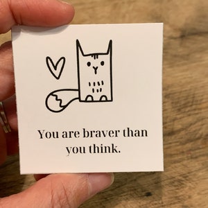 May include: A white card with a black and white illustration of a cat with a heart above it. The text on the card reads "You are braver than you think."