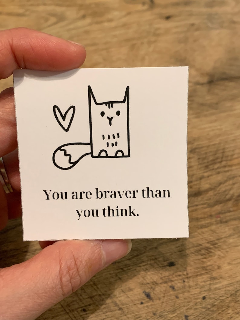 May include: A white card with a black and white illustration of a cat with a heart above it. The text on the card reads "You are braver than you think."