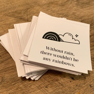 May include: A stack of white cards with a black and white illustration of a rainbow and a cloud with a star. The text on the card reads "Without rain, there wouldn't be any rainbows."