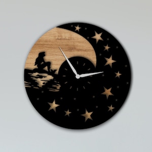 May include: A black and wood wall clock with a crescent moon and stars design. The clock features a silhouette of a mermaid sitting on a rock.