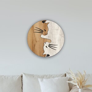 May include: A wooden round wall decor with a yin yang design featuring two cats facing each other. The left side of the circle is brown wood with a black silhouette of a cat with brown fur. The right side of the circle is white wood with a black silhouette of a cat with white fur.
