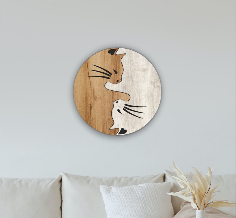 May include: A wooden round wall decor with a yin yang design featuring two cats facing each other. The left side of the circle is brown wood with a black silhouette of a cat with brown fur. The right side of the circle is white wood with a black silhouette of a cat with white fur.