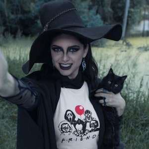 May include: A woman wearing a black witch hat and a white t-shirt with a graphic of horror movie characters. The shirt says "F.R.I.E.N.D." and features a red balloon. She is holding a black cat.