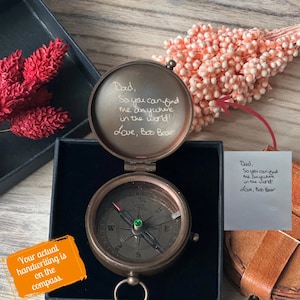 May include: A compass with a handwritten message on the lid that reads "Dad, So you can find me anywhere in the world! Love, Boo Bear". The compass is open and the needle is pointing north. The compass is in a box with a brown leather pouch.