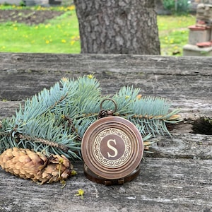 May include: A vintage brass compass featuring the letter "S" in the center, displayed on a weathered wooden surface with pine needles and a pine cone. The compass has an ornate design around the letter, creating a rustic, antique aesthetic.