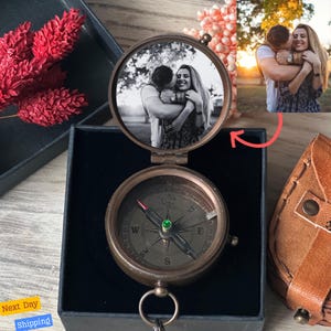 May include: A brass compass with a photo insert on the top. The compass has a black face with white markings and a green jewel in the center. The photo insert shows a couple embracing in a black and white photo.