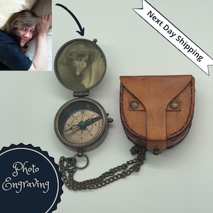 May include: A brass compass with a photo insert, a leather pouch, and a chain. The compass has a green needle and a white face with black markings. The photo insert shows a person's face. The pouch is brown and has two metal studs. The chain is brass and connects the compass to the pouch.