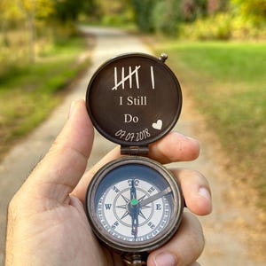 May include: A compass with a heart engraved on the lid. The lid is open and says "I Still Do" with a date. The compass is in a hand.