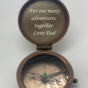 May include: A vintage brass compass with the lid open, revealing the inscription "For our many adventures together Love Dad." The compass features a detailed dial with directional markings and a needle with a green jewel. The compass is on a white background.
