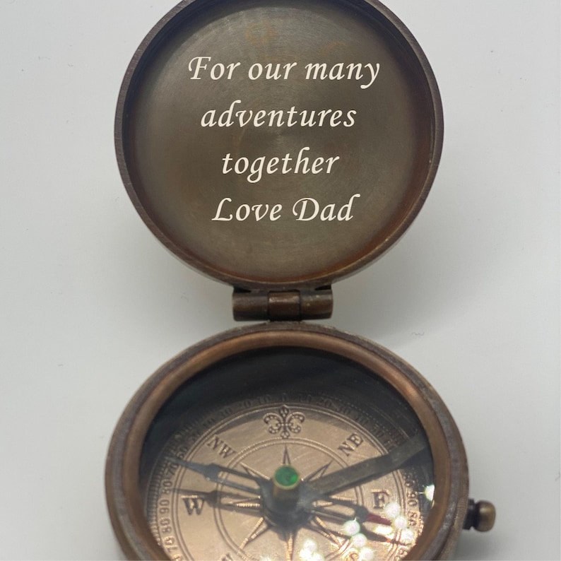 May include: A vintage brass compass with the lid open, revealing the inscription "For our many adventures together Love Dad." The compass features a detailed dial with directional markings and a needle with a green jewel. The compass is on a white background.