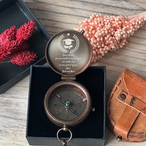 May include: An open antique brass compass with a graduation cap and scroll engraving. The compass is in a black box, with text that reads "Dear David, you are worth every sacrifice we made, and we are proud of you. Love, Dad and Mom."